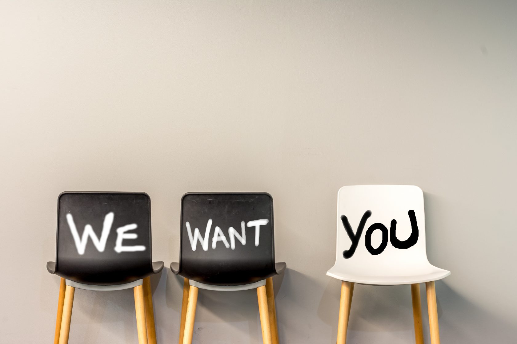 Job recruiting advertisement represented by 'WE WANT YOU' texts on the chairs. One chair is colored differently and labeled 'YOU' to represent the hiring position to be recruited and filled.